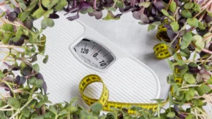 White dial bathroom scale reading zero kg with a coiled yellow tape measure, framed by green and purple radish seedlings