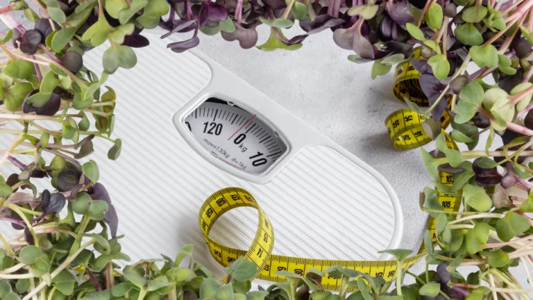 White dial bathroom scale reading zero kg with a coiled yellow tape measure, framed by green and purple radish seedlings