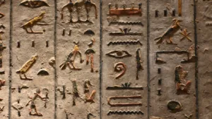 Carved ancient Egyptian hieroglyphs painted in gold and ochre — birds, eyes and figures — in vertical columns on weathered stone