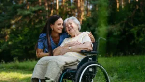 Smiling carer in blue scrubs leans down to embrace a laughing white-haired woman seated in a wheelchair in a sunlit park
