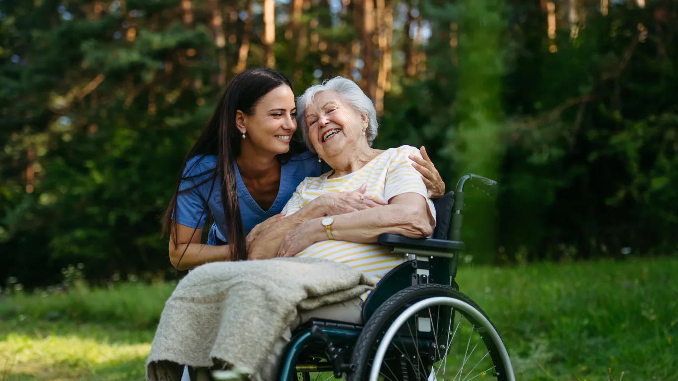 Smiling carer in blue scrubs leans down to embrace a laughing white-haired woman seated in a wheelchair in a sunlit park