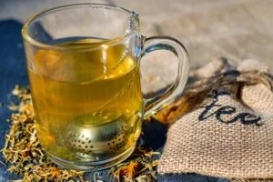 Glass mug of golden herbal tea with a metal infuser ball, loose dried flowers on slate beside a burlap sack marked "tea"