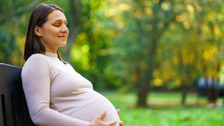 Pregnant woman sitting on a park bench with her eyes closed and a faint smile, both hands resting on her belly, sunlit green trees blurred behind her
