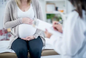 Pregnant woman in a grey cardigan sits on an examination couch cradling her bump while a clinician in a white coat writes on a clipboard in the foreground