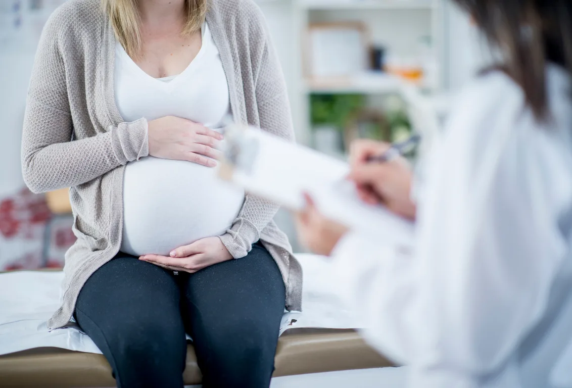Pregnant woman in a grey cardigan sits on an examination couch cradling her bump while a clinician in a white coat writes on a clipboard in the foreground