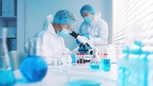 Two technicians in white protective suits, hairnets, masks and goggles work in a blue-lit laboratory; one leans into a microscope beside a rack of red-capped blood tubes and blue-filled flasks
