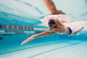 Underwater view of a swimmer in a dark cap and goggles mid front-crawl stroke, arm extended, trailing bubbles beside a red and white lane rope in a blue pool