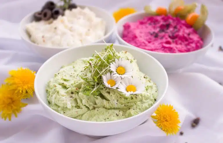 Three white bowls of dips: a green herb dip topped with cress shoots and daisies, a white olive dip, and bright pink beetroot dip