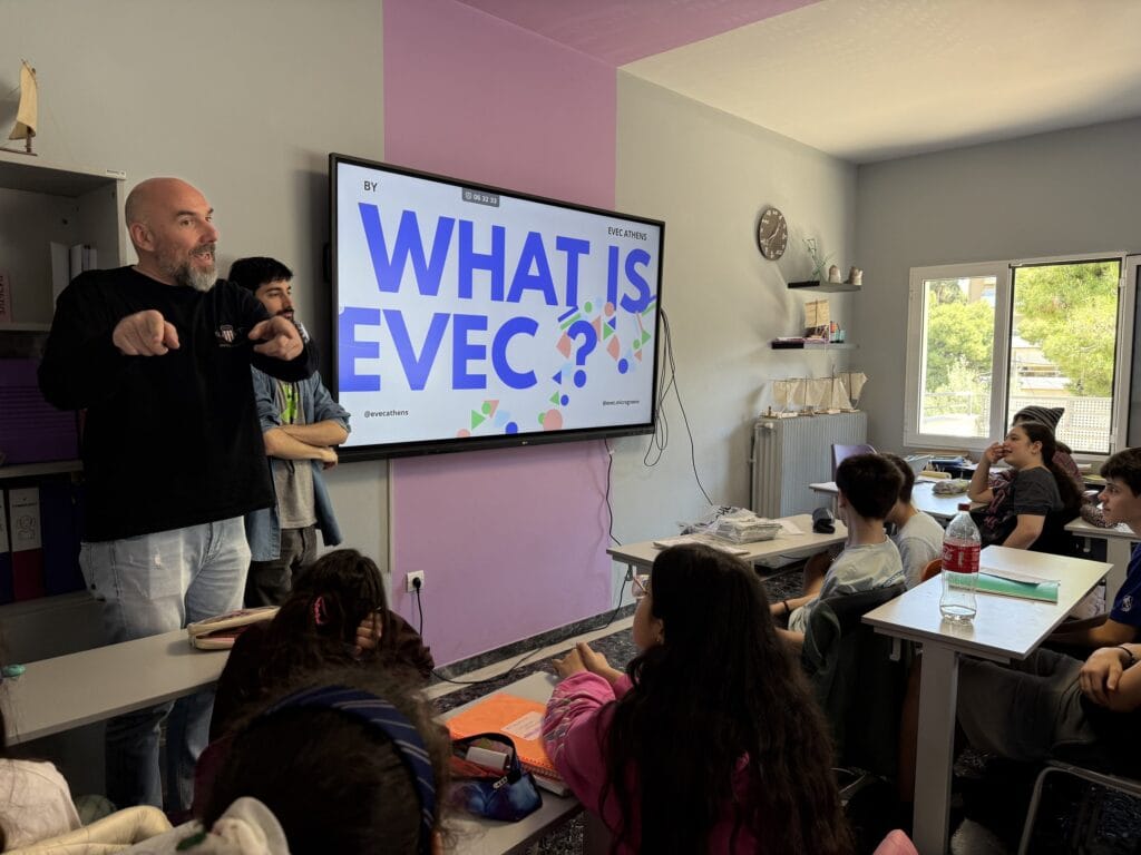 A trainer presenting an introductory EVEC Athens slide to a class of school students seated at their desks.